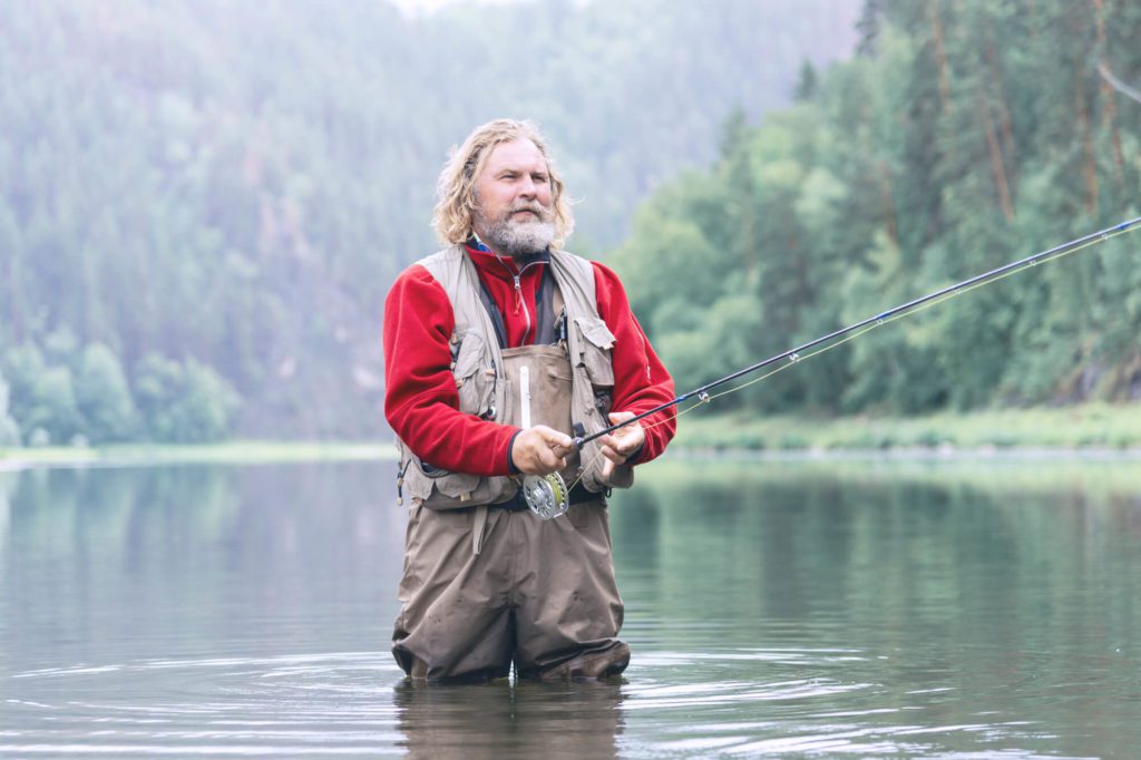 bearded fisherman in a red jacket and overalls stands in the water with a fishing rod.